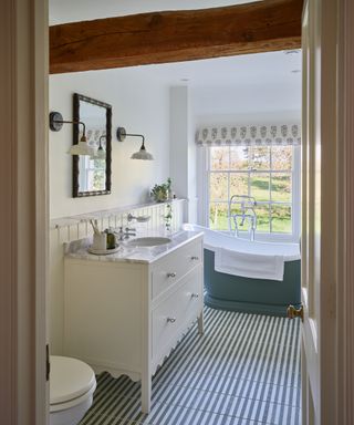 green freestanding bath below window in bathroom with green and white tiled floor, white vanity unit with inset sink, wooden beams on celing, roman blind at window and wall mounted lights and mirror above sink