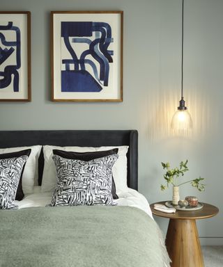 bedroom with green walls, bed with hanging black and glass pendant to side above wooden bedside table with two blue and whte modern art prints above bed