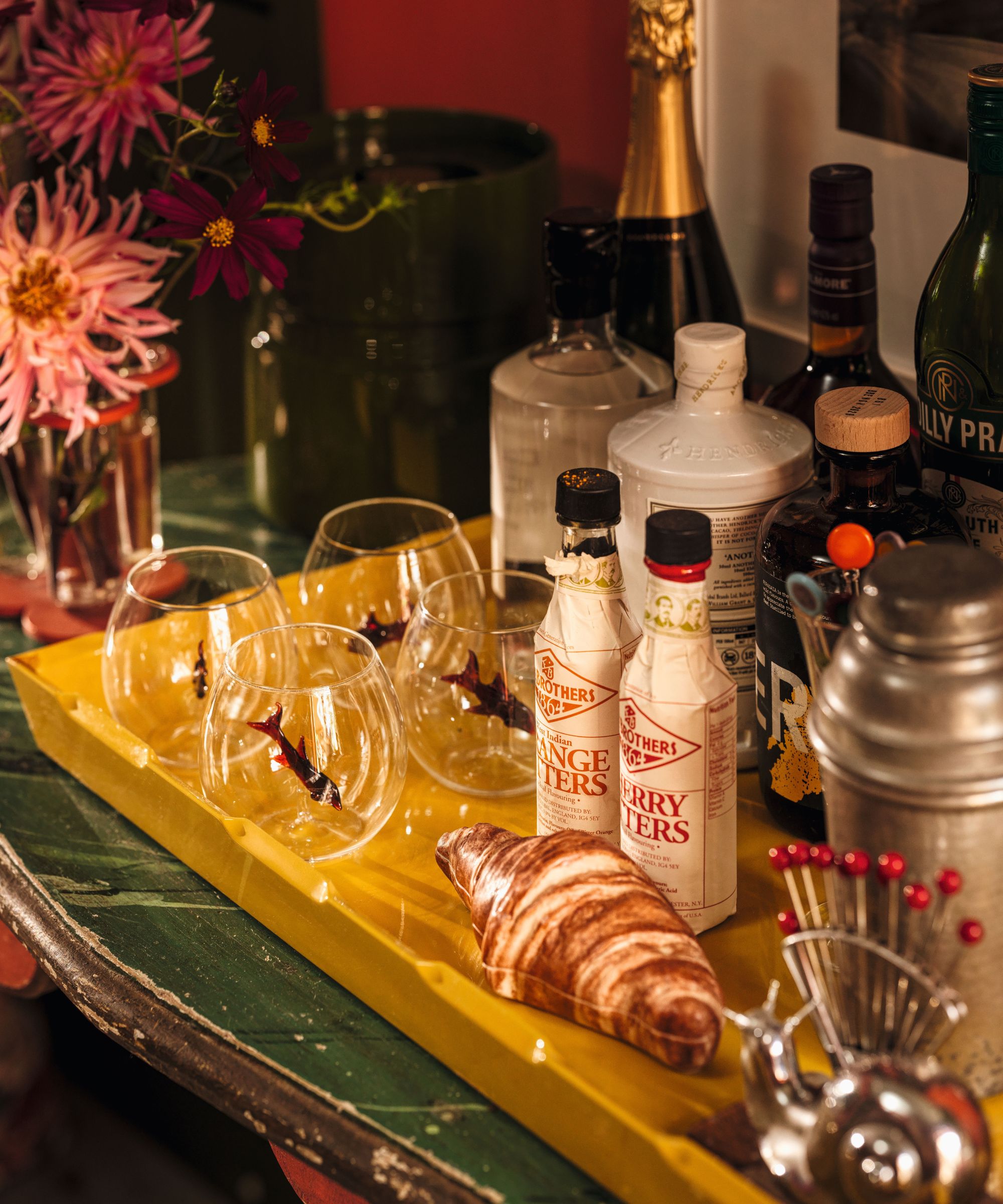 Yellow tray with fish-embellished cocktail glasses, Fee Brothers bitters, gin, a croissant-shaped accessory, cocktail shaker, and pink dahlias.
