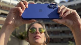 Apple iPhone 16 Plus held up in two hands to take a photo by a person wearing sunglasses