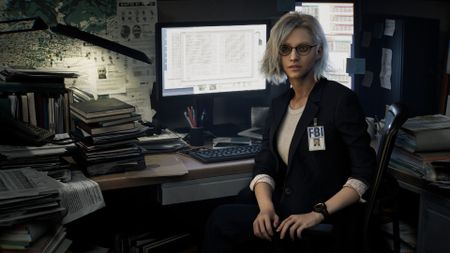 Resident Evil Requiem screenshot of Grace Ashcroft, who has white short hair and glasses and wears a black blazer with an FBI badge, sitting at a desk in front of a PC monitor