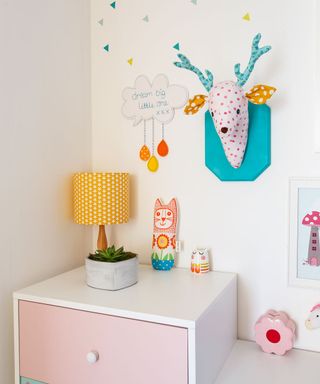childrens room with white wall with painting and stickers and cabinet with lamp with yellow shade and toys