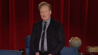 Conan standing in front of a red curtain and props during his final farewell on Conan, TBS. He has on a suit, tie, and tie clip. The suit is unbuttoned.