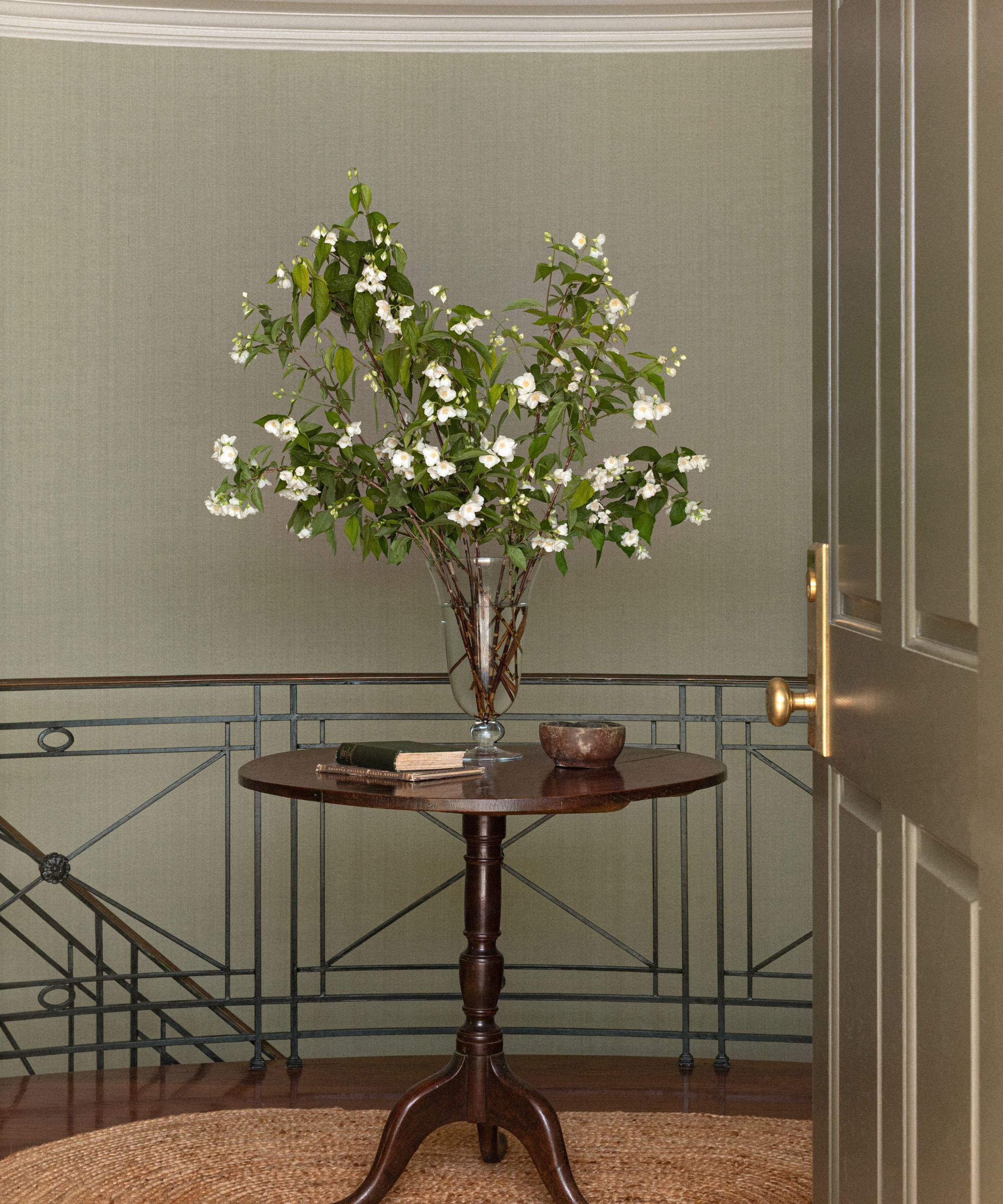 an oval entryway with black stair railings, green grasscloth wallpaper, with an antique table with a vase of flowers sat on a woven rug