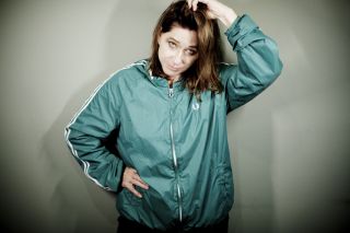 Kim Deal stands in front of a grey background with her hand on her head. She is wearing a green zip up jacket