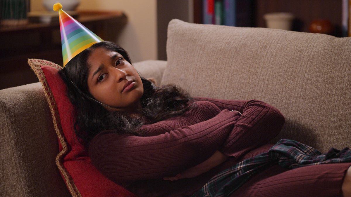 Devi (Maitreyi Ramakrishnan) lies on a sofa wearing a rainbow party hat in Never Have I Ever season 3.