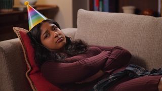 Devi (Maitreyi Ramakrishnan) lies on a sofa wearing a rainbow party hat in Never Have I Ever season 3.