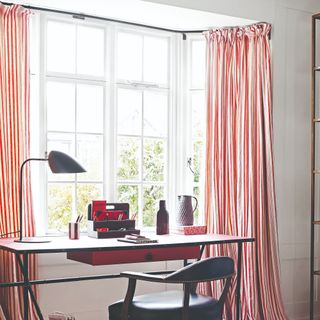 A home office with the desk facing a window adorned with striped curtains and a modern desk lamp on the desk