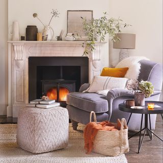 Grey velvet armchair with geo print footstool in front of a wood burner, with vases lined up along a white marble fireplace