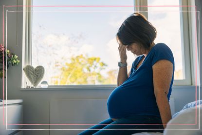 A woman sits on a bed by a window while experiencing a pregnancy migraine