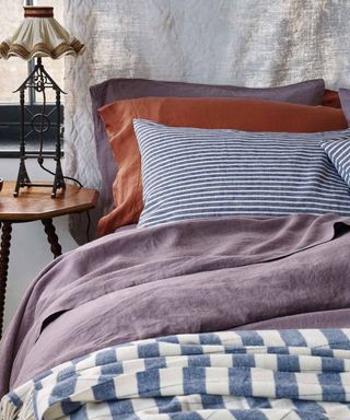 Elderberry, rust, and blue and white striped bedding on a bed against a gray curtain.