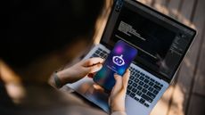 An over-the-shoulder shot of a worker on a laptop, while holding a phone showing an AI copilot logo.