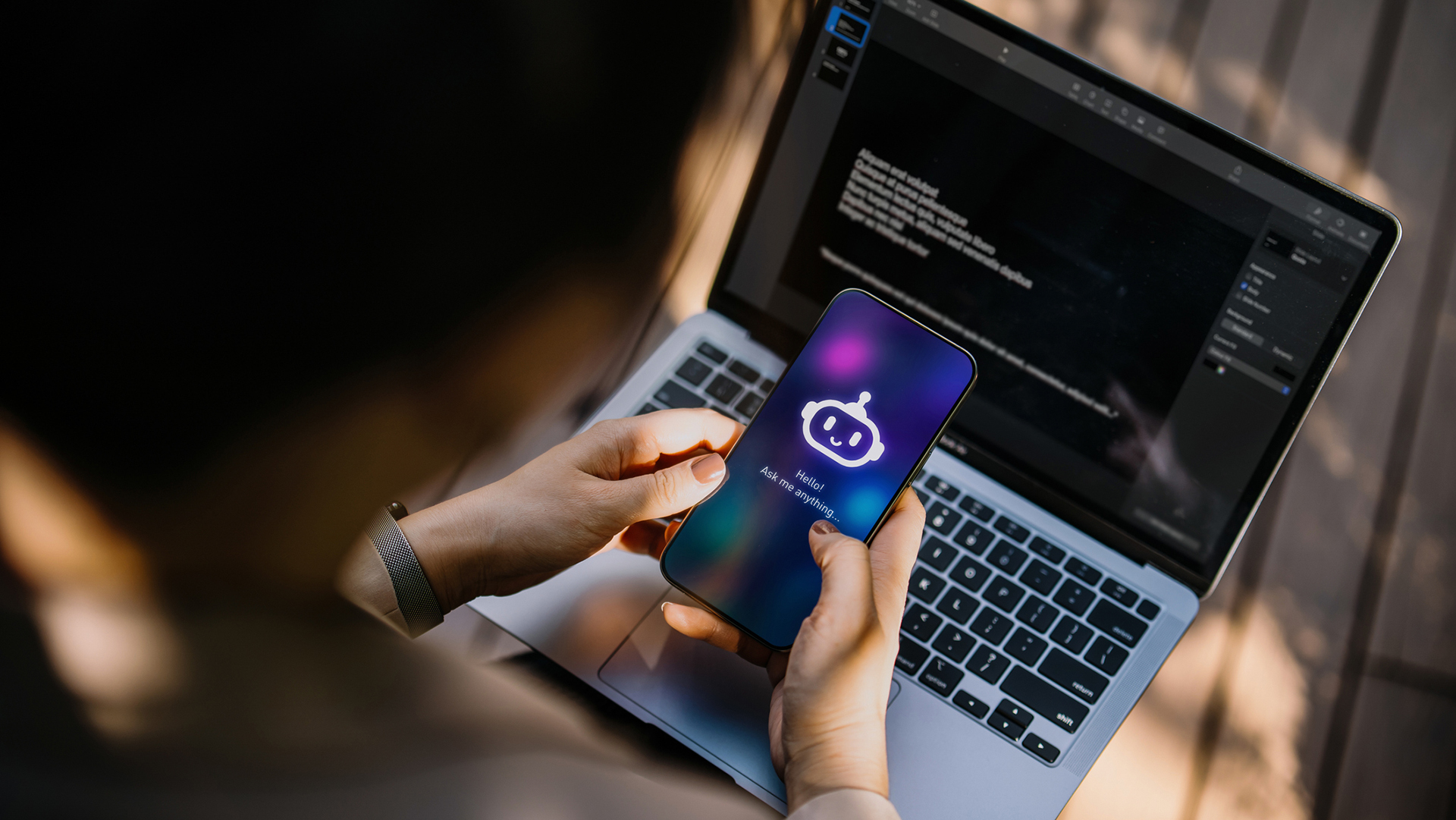 An over-the-shoulder shot of a worker on a laptop, while holding a phone showing an AI copilot logo.