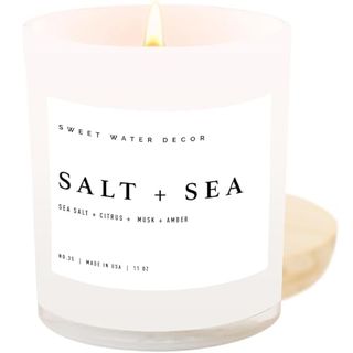 A frosted white glass jar with a white candle inside lit with a flame. A white label with 'Salt + Sea' printed on the front. 