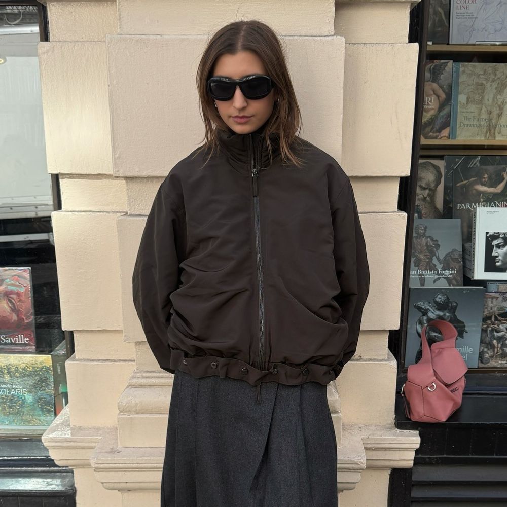 Influencer @poppyalmond poses for a phot wearing oversized sunglasses and a brown bomber jacket.