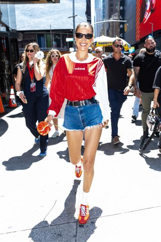 Bella Hadid walks through times square wearing an adidas soccer jersey jean shorts and adidas sneakers