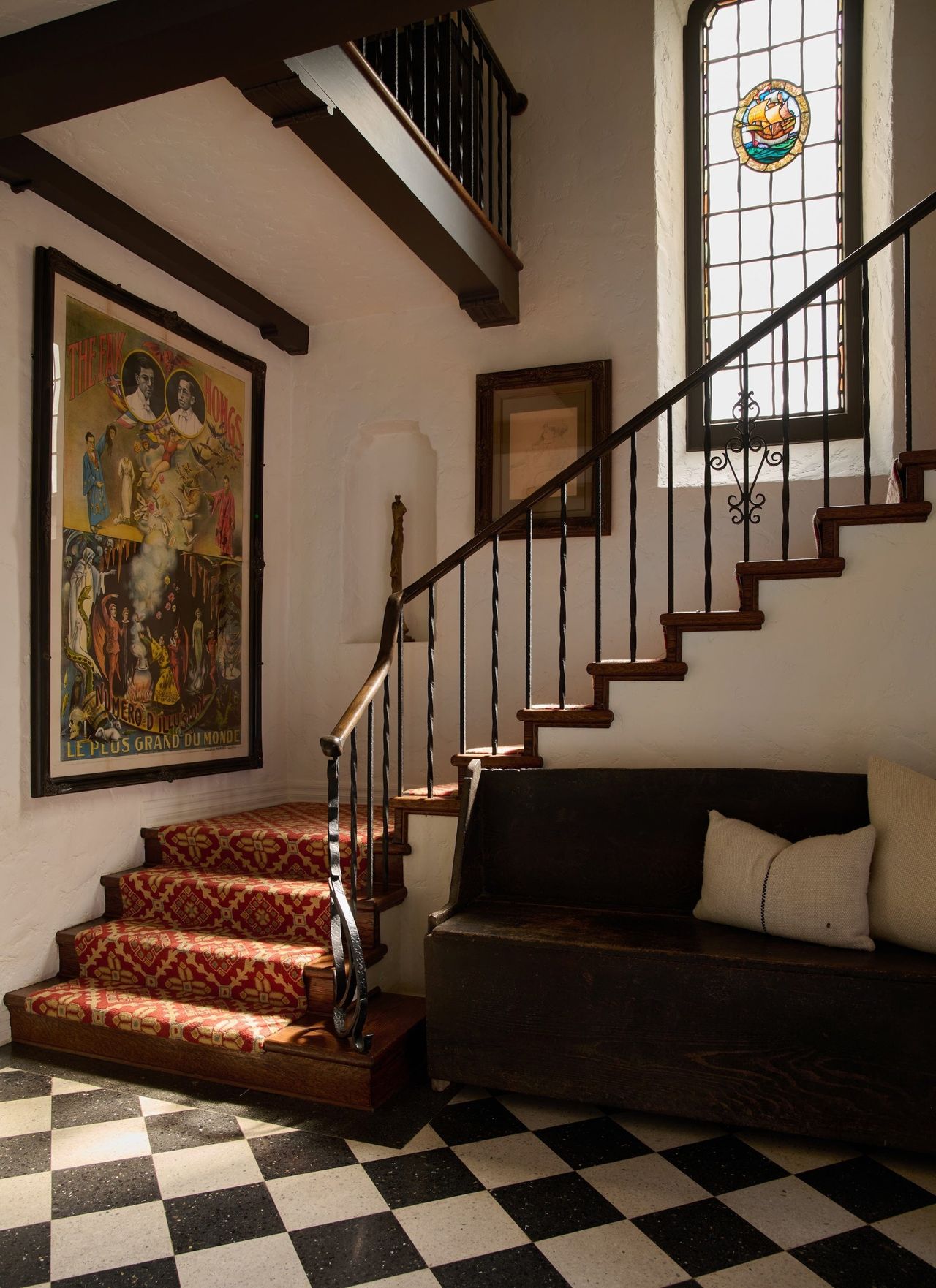 checkerboard black and white tiled entryway with winding staircase with red and yellow patterned runner. long window above stairs with stain glass detailing