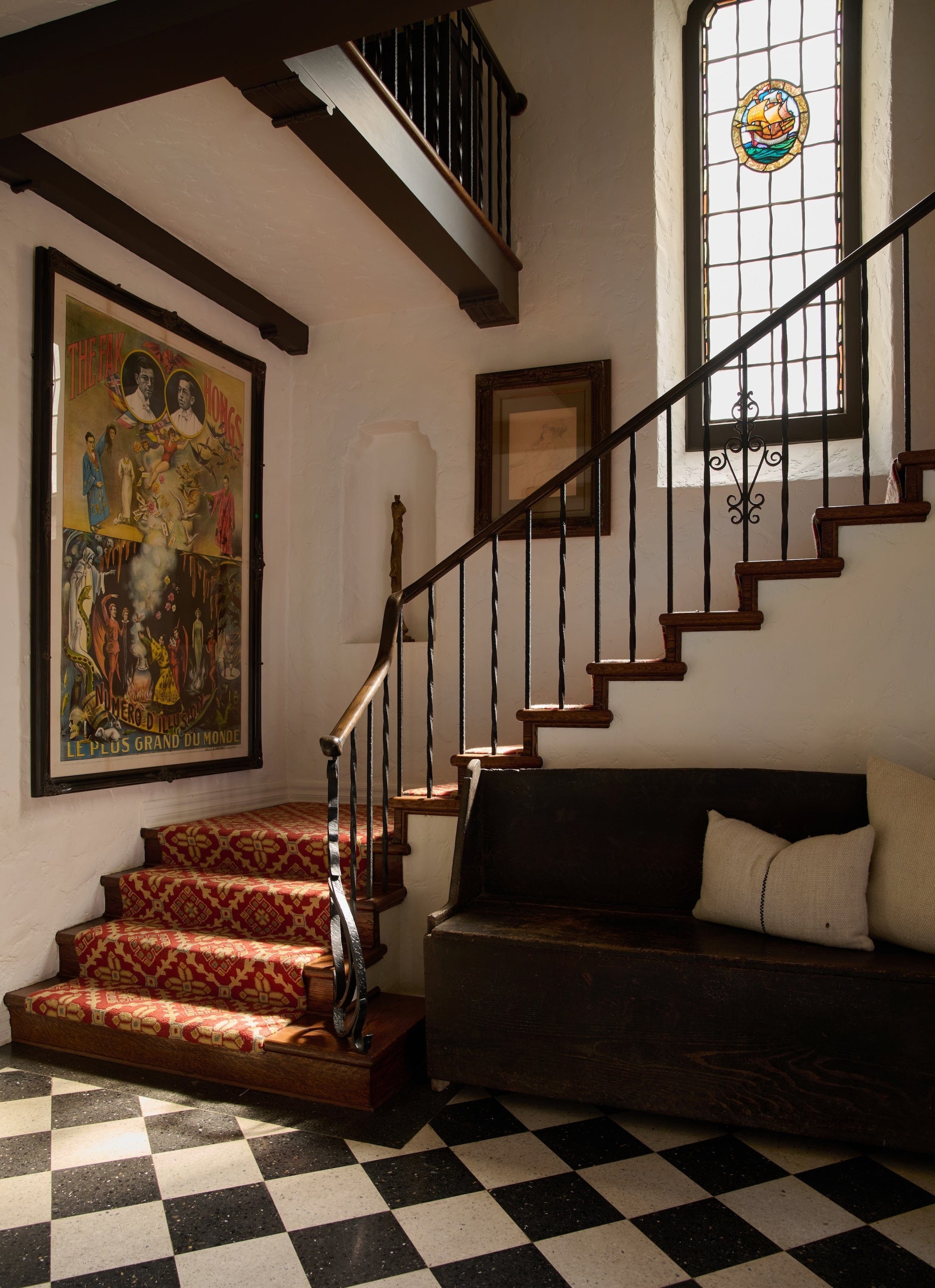 checkerboard black and white tiled entryway with winding staircase with red and yellow patterned runner. long window above stairs with stain glass detailing 