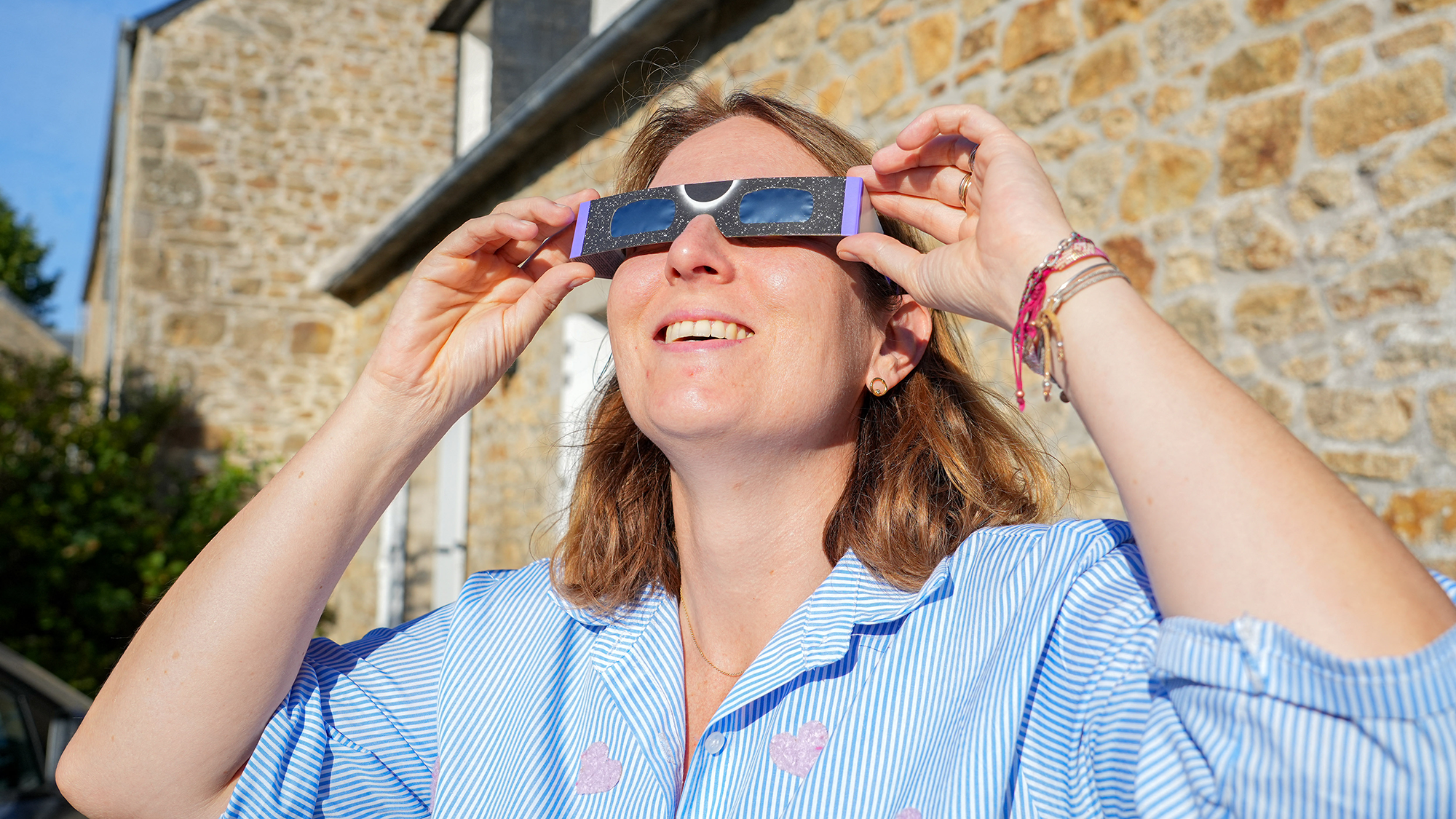 A woman with short blond hair wears eclipse glasses and stares upward