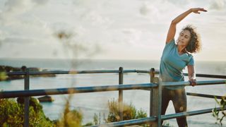 Woman cooling down after running by stretching, arm up over her head, looking out at sea