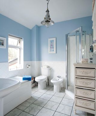 Small bathroom with white tiles on bottom third of walls and light blue pain above, with a white bath, toilet, shower suite, and ornate, tall drawer cabinet for storage
