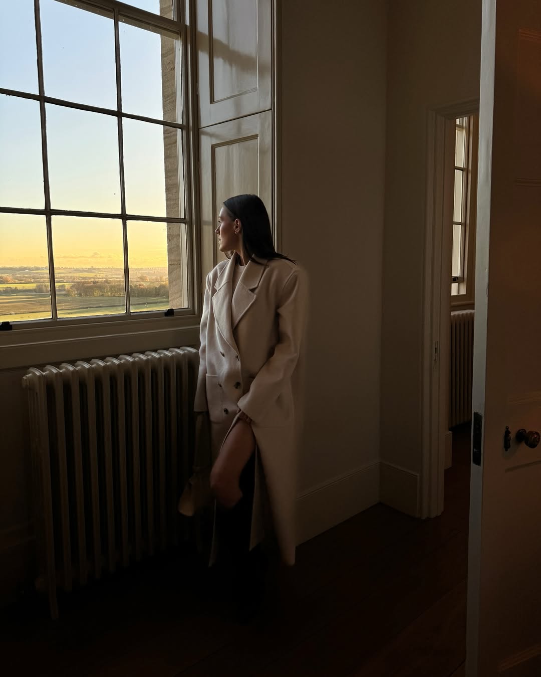 Emma Leger poses near a window in an oversized cream coat while on a trip to The Cotswolds