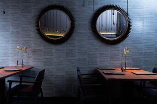 Two wooden tables, each with a minimalist floral centerpiece, stand underneath two black mirrors in a dimly lit restaurant.