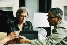 Two older women meet at a table, discussing something over a computer tablet.