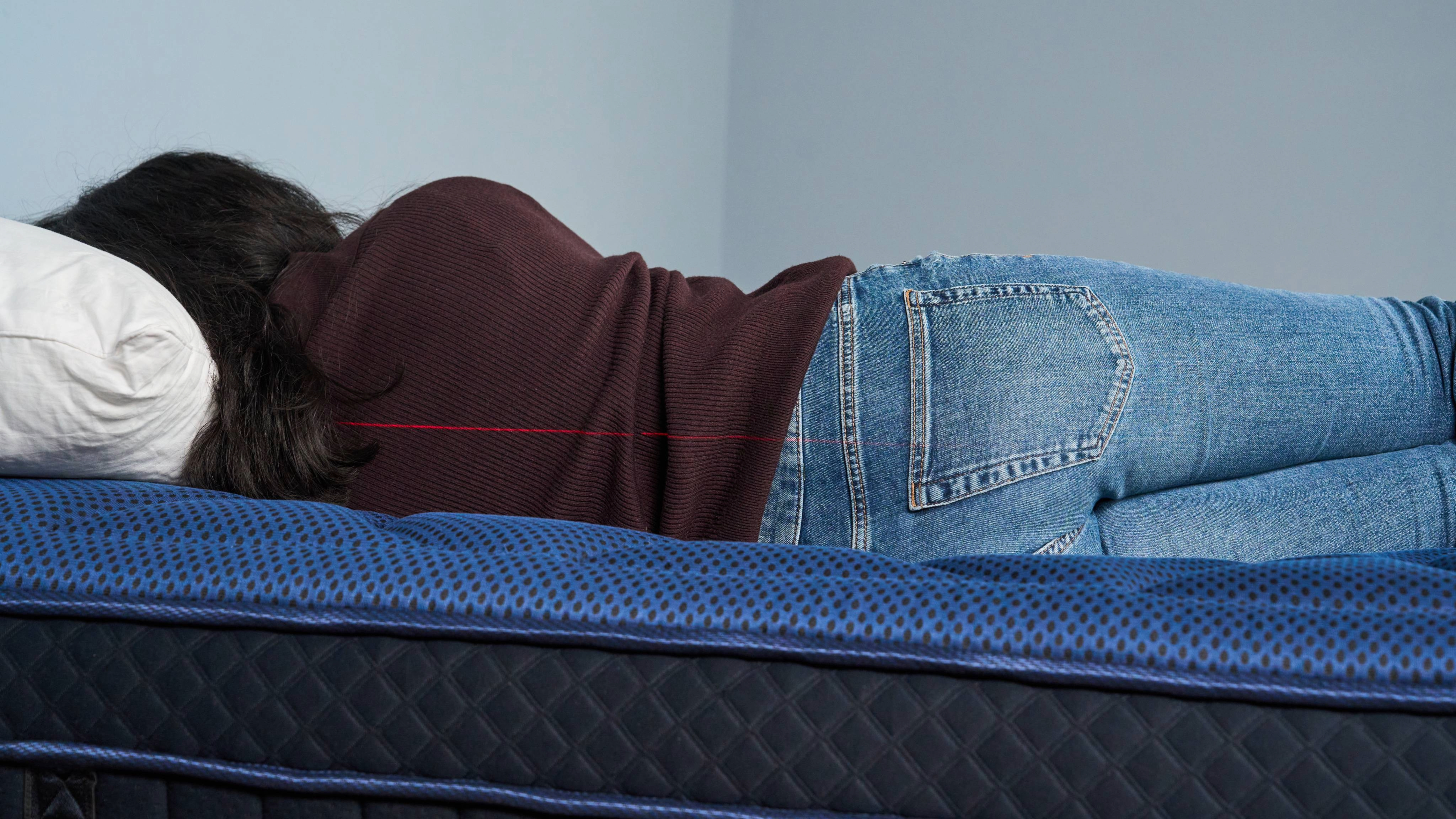 The image shows a dark haired mattress tester lying on her side on a dark blue mattress, with her back to the camera. Along her back, a laser level marks how deeply her spine sinks into the bed.