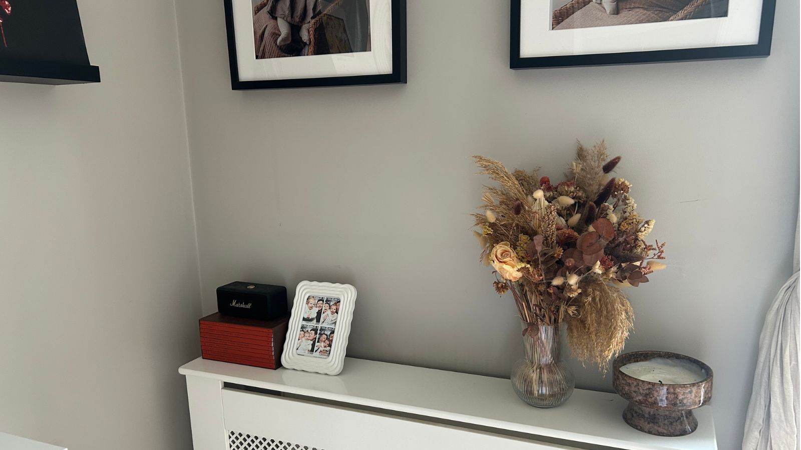 Corner of living room with radiator shelf, frames on wall and dried flower bouquet