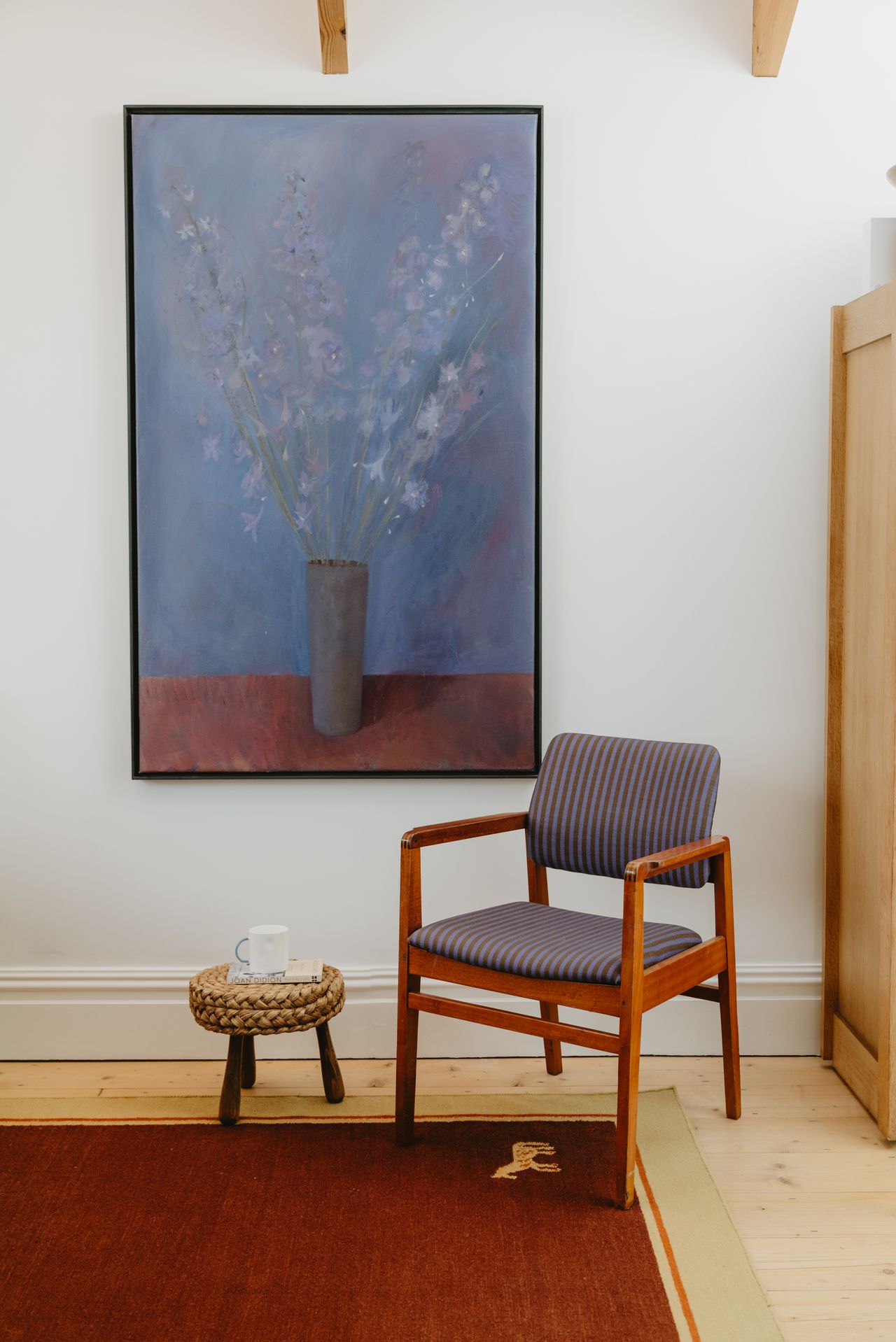 Image of a dark periwinkle stripe and wooden accent chair on a red rug in a white room. There is a blue and red painting of a vase of flowers on the wall.