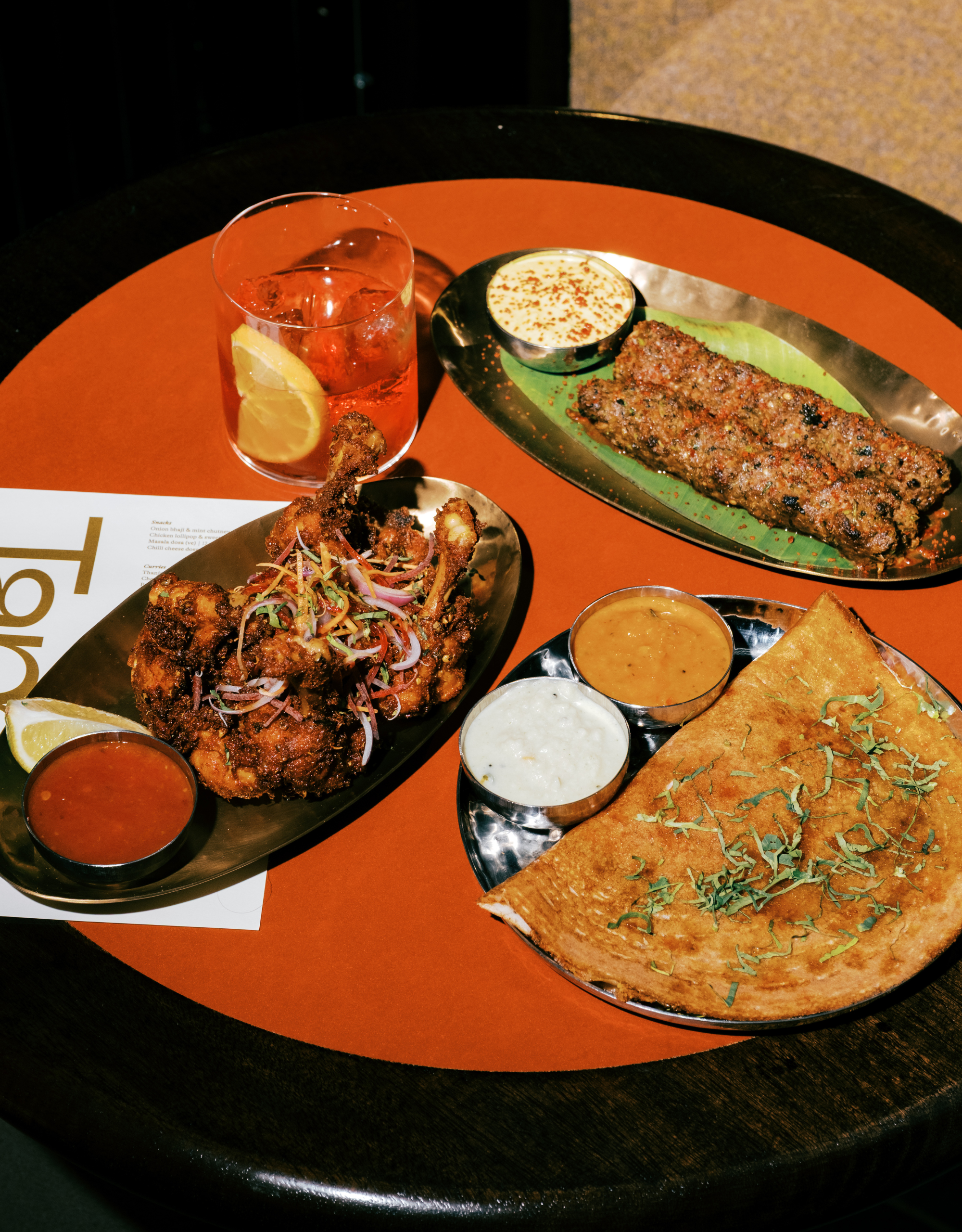 An orange table at Tamila, London with steel dinnerware serving chicken fry, dosa with chutney and sambar, and seekh kebabs beside a drink