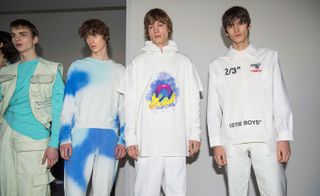 Four male models wearing looks from Off-White's collection. One model is wearing a blue top, cream sleeveless piece with pockets and cream combat trousers. Another model is wearing a white and blue tracksuit. The third model is wearing a white hooded top with colourful design at the front and white trousers. And the fourth model is wearing a white shirt with black wording and white trousers