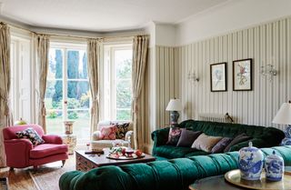 Green sofas in living room with striped wallpaper and pink and cream armchairs vintage wood coffee table and big bay window
