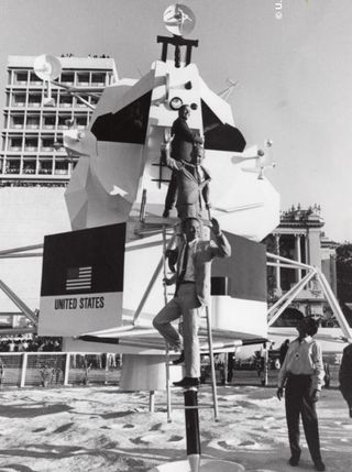 three men in suits climb on a mockup of a lunar lander