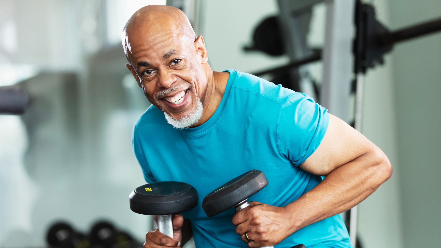 A man holds a pair of heavy dumbbells and smiles at the camera.