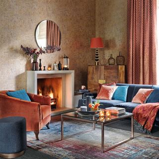 Living room with patterned wallpaper, blue sofa, red chair, fireplace, glass top table and patterned rug.