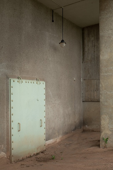 A heavy pale green steel door studded with rivets and fitted with handles stands against a bare concrete wall, beneath a single pendant lamp hanging from the ceiling, with sand and weeds on the floor.