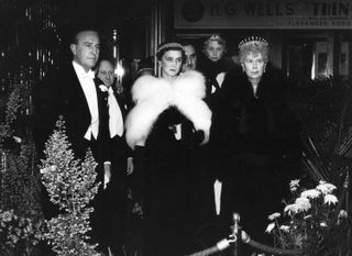 Queen Mary wearing a tiara and black coat standing next to Duchess Marina of Kent wearing a white fur wrap and tiara next to a man in a tux at a theater performance