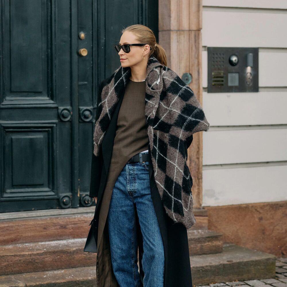 Woman poses while wearing jeans, snake print boots, an asymmetrical skirt with long black coat and plaid sweater over their shoulder.