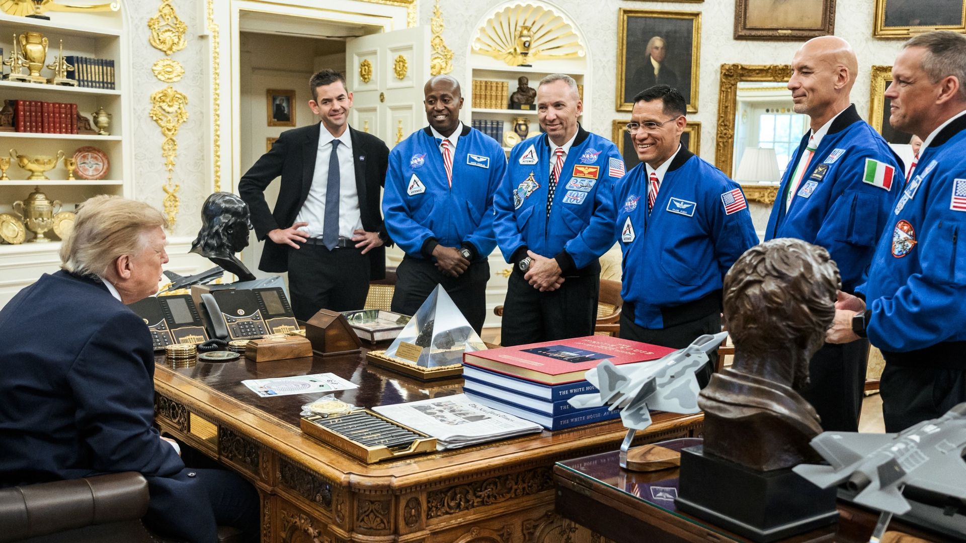 a man seating at a desk in a fancy room talks to one man in a dark suit and five male astronauts in blue flight jackets