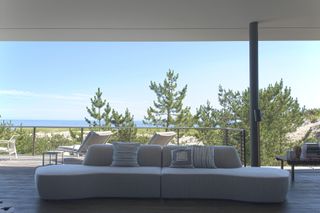 A gray sofa sits at the center. Behind it, we see the deck of the beach house and the sea in the distance.