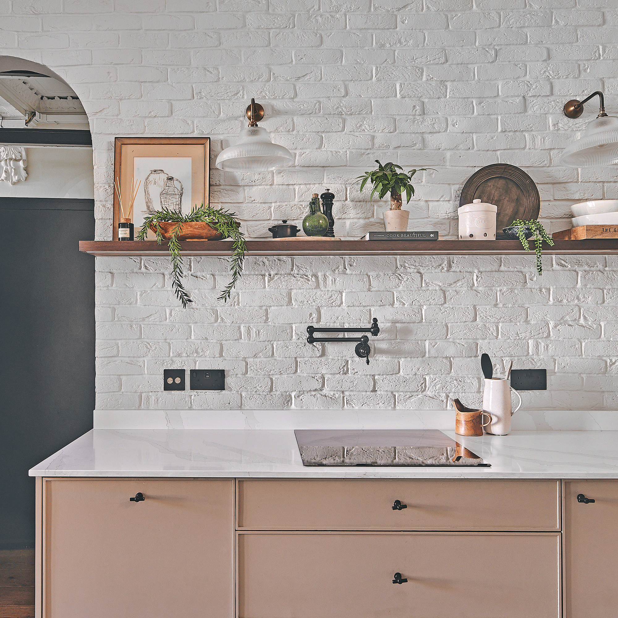a kitchen with white painted exposed brick walls and beige modern cabinets and an induction hob with open shelving above it