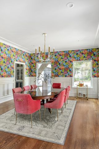 dining room with white wainscoting, floral wallpaper above, red chairs, rug, mahogany table, wooden floor, bar cart