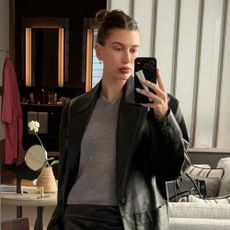 Hailey Bieber takes a mirror selfie wearing a leather knee-length pencil skirt with knee-high pointed-toe boots, a grey v-neck jumper and a black leather blazer.