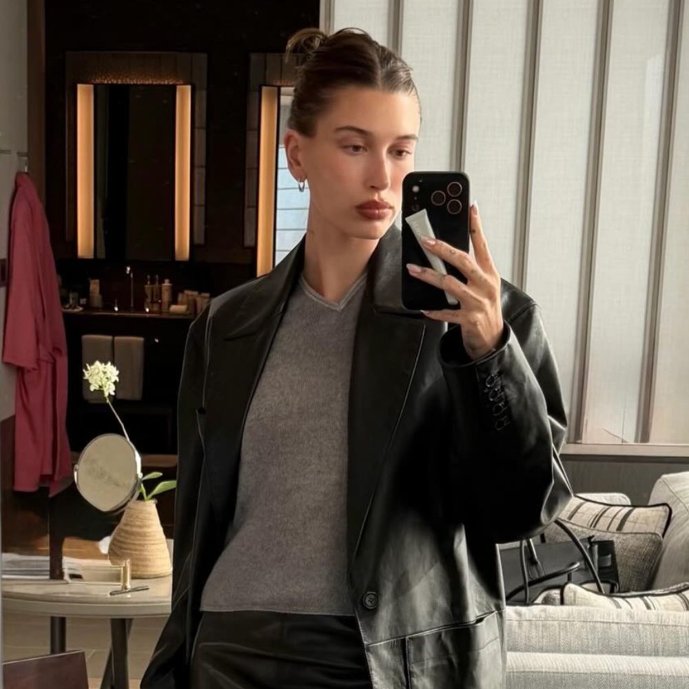 Hailey Bieber takes a mirror selfie wearing a leather knee-length pencil skirt with knee-high pointed-toe boots, a grey v-neck jumper and a black leather blazer.