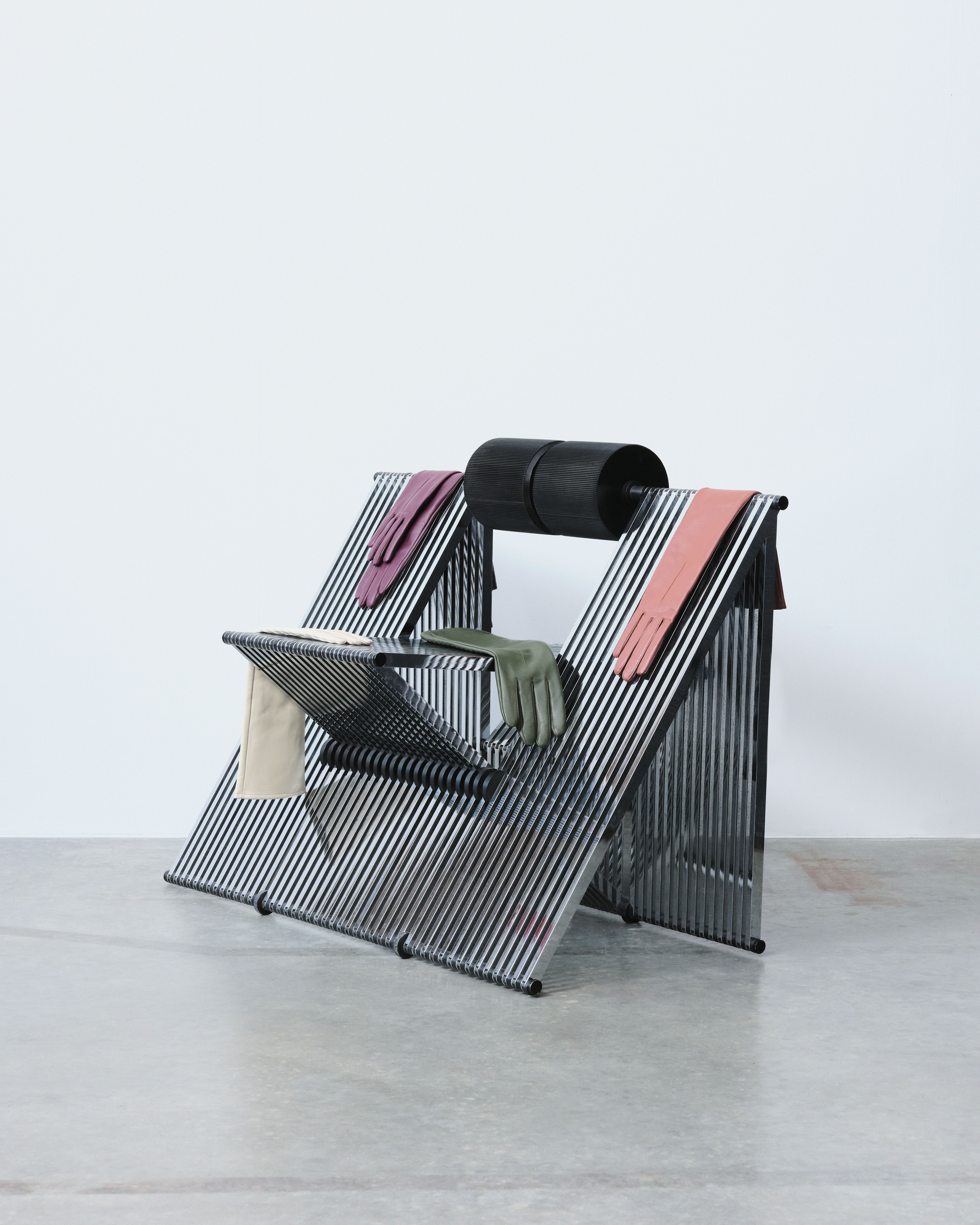 A chrome made, foldable elevated sculptural chair with a leather roll-like back and a multi-triangular structure is covered in colorful leather gloves while sitting in an industrial site.