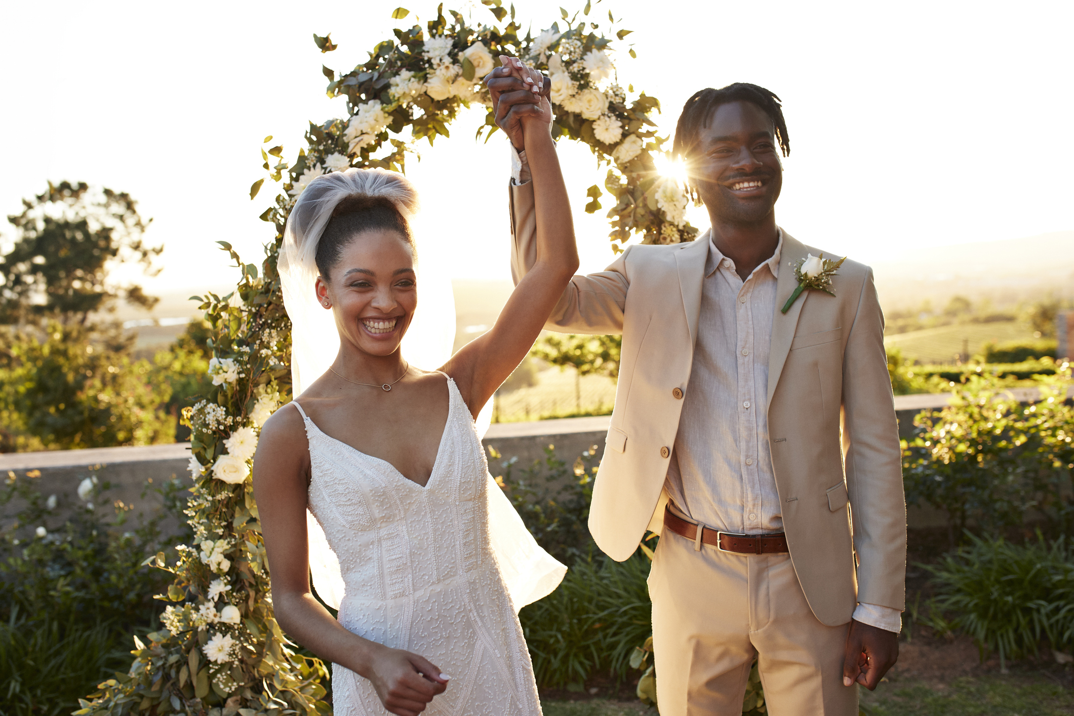 Bride and groom hold hands aloft backlit against the sun on their wedding day