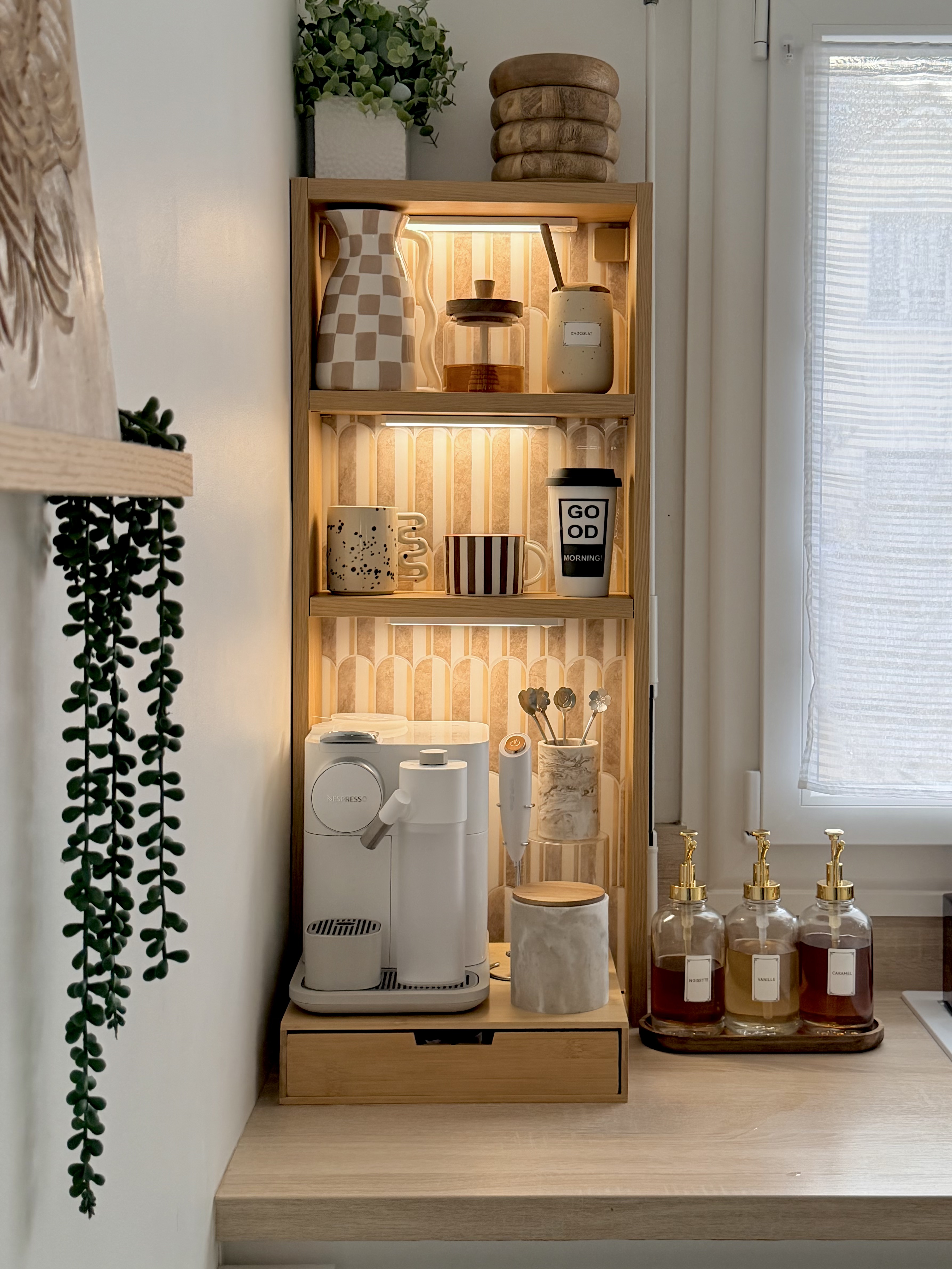 Image of the IKEA HAAGAN shelf being used as a coffee station in a white kitchen. The shelf is equipped with a coffee maker and all of the necessary coffee accessories. The back of the shelving has wallpaper and there are led strip lights.