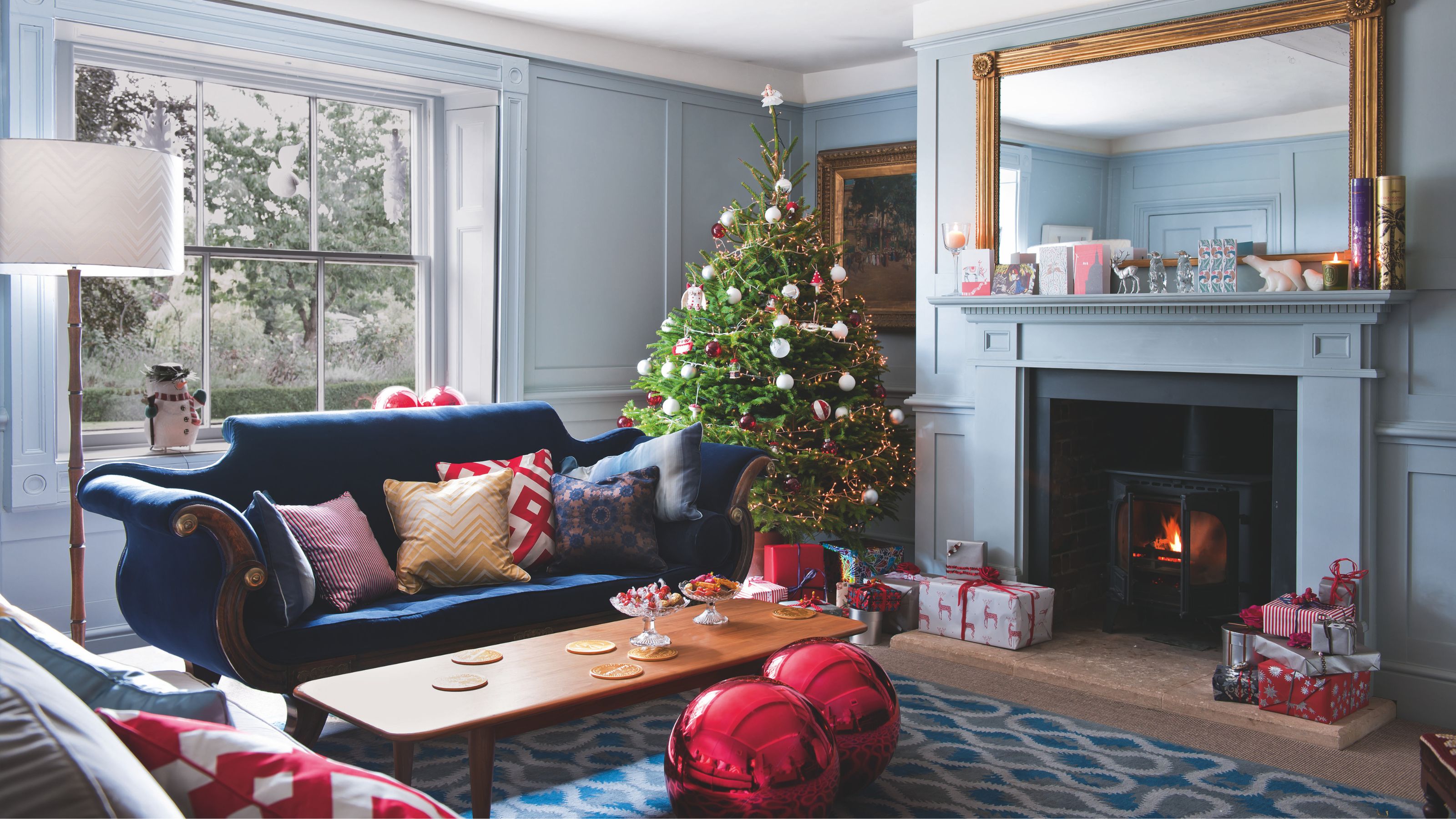 Festive living room with blue wall panelling, a dark blue sofa covered in cushions, an open fireplace and a christmas tree in the corner